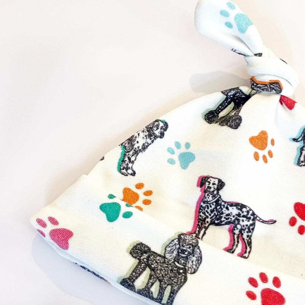 A close-up view of the baby hat showcasing the dog illustrations and vibrant paw prints on a soft white fabric background The print includes 7 adorable dog breeds sketched in black on a white fabric background and accented with colorful paw prints and highlights in yellow, orange, green, turquoise & light blue.