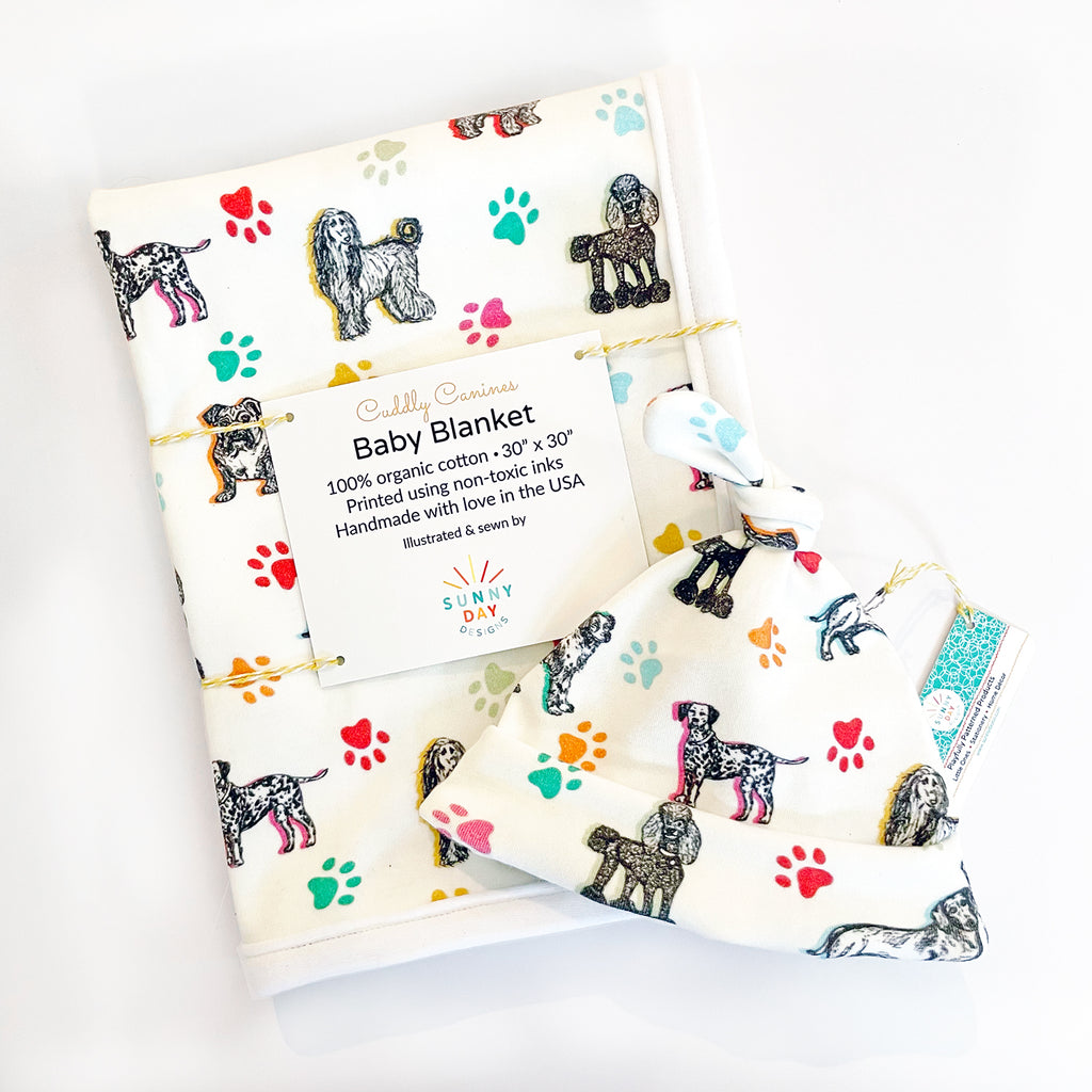 A super soft organic cotton dog-printed baby blanket and newborn hat gift set by Sunny Day Design is shown on a white background. The print on both of these Cuddly Canines printed  baby gifts includes 7 adorable dog breeds sketched in black on a white fabric background and accented with colorful paw prints and highlights in yellow, orange, green, turquoise & light blue.