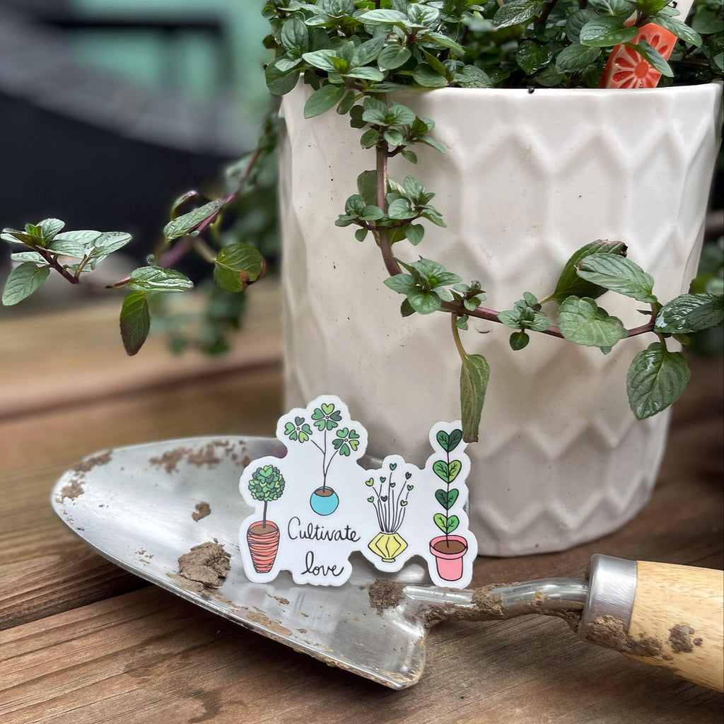 A gardening-themed Cultivate Love Vinyl Sticker designed by Sunny Day Designs is shown resting on a garden trowel next to a potted Chocolate Mint plant inside a white ceramic planter. Each cute plant-themed sticker features hand lettered "Cultivate Love" text with 4 potted plants with heart-shaped leaves planted inside assorted colorful planters.