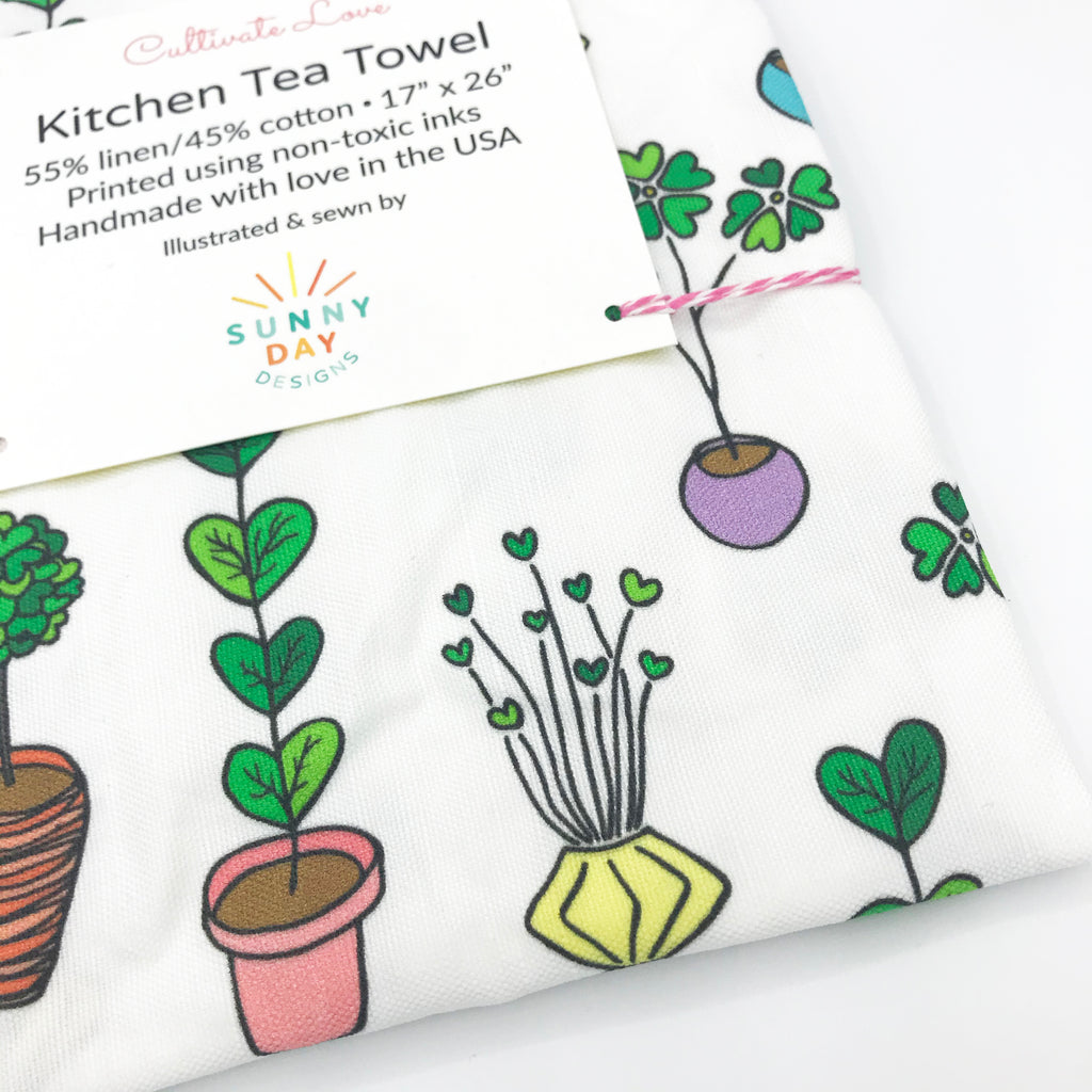 A close-up view of an indoor plant-themed printed kitchen tea towel, which features a variety of green leafy plants with heart-shaped foliage inside of colorful pots on a white background.