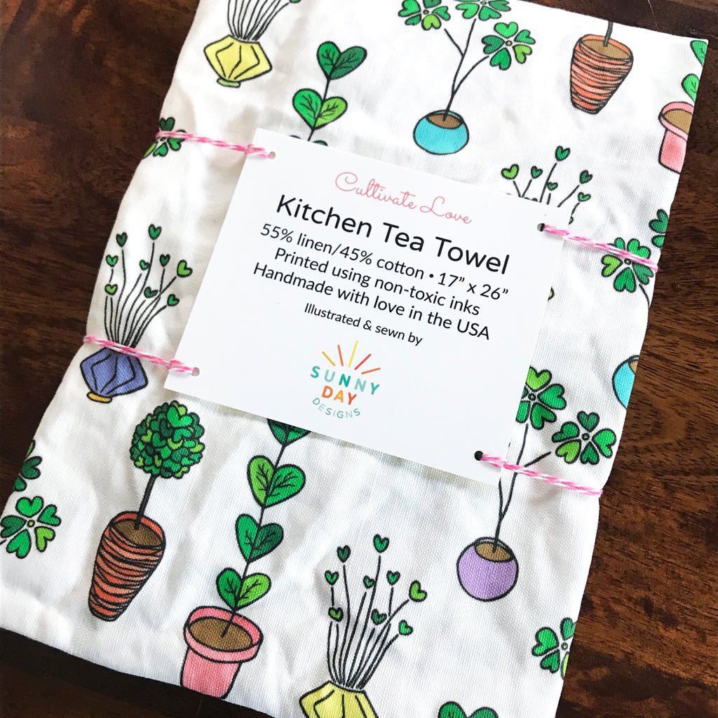 A handmade, colorfully-printed linen/cotton plant-themed kitchen dish towel by Sunny Day Designs is shown on top of a dark brown wooden table. This whimsically printed tea towel is joyfully in made in the USA and makes a perfect gift for Valentine's Day.