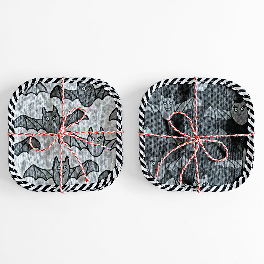 Set of Halloween-themed cocktail napkins by Sunny Day Designs featuring a playful bat pattern. Two sets of napkins are shown side by side in light and dark colorways, each tied with red and white twine and bordered with black and white striped edging.