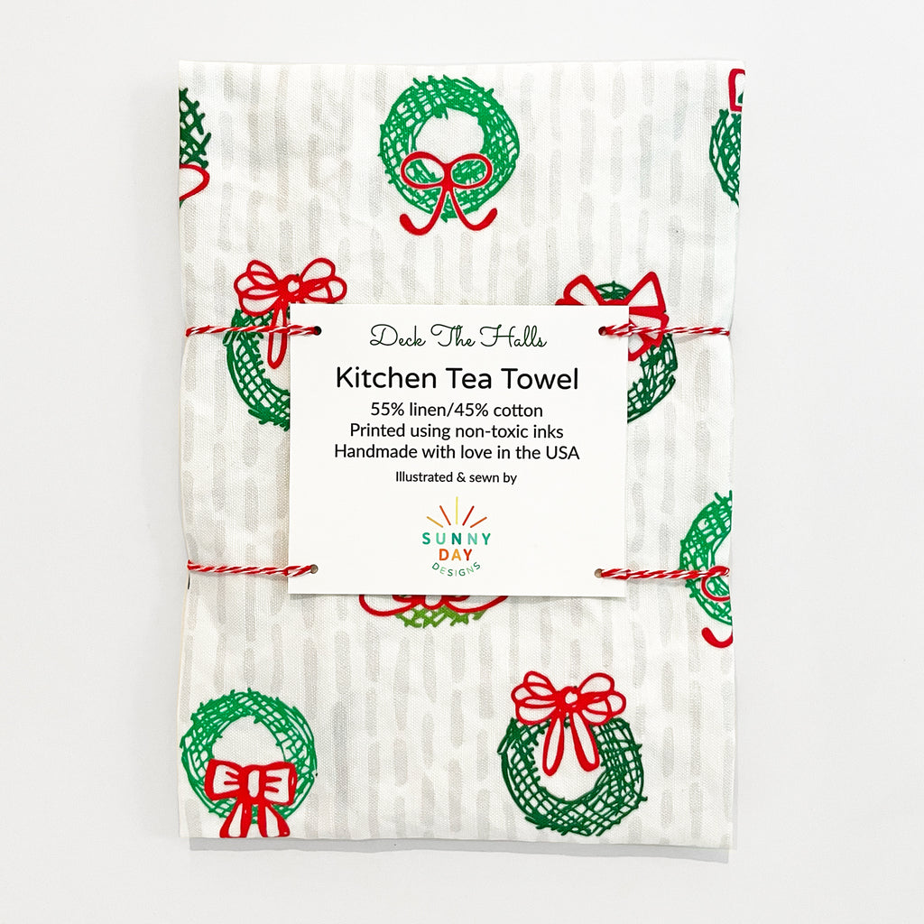 Deck the Halls holiday kitchen tea towel with green wreaths and red bows pattern, made of 55% linen and 45% cotton. The towel is packaged with a label that reads 'Deck The Halls Kitchen Tea Towel,' featuring details about non-toxic inks and being handmade in the USA by Sunny Day Designs.