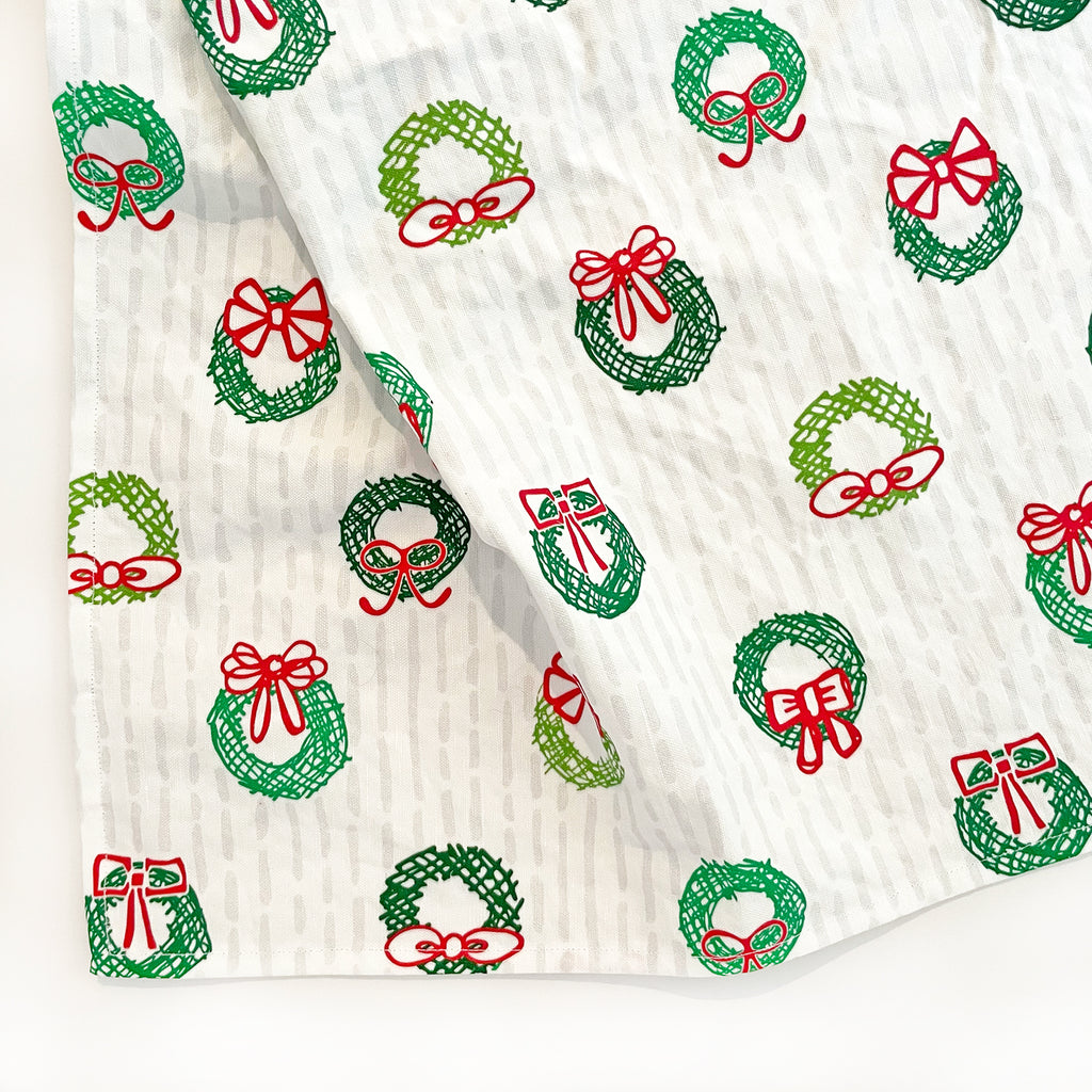 A new print for the 2024 holiday season, our Christmas wreath themed "Deck The Halls" printed kitchen towel is shown un-packaged with the fabric draped on a white background to show the print design. Each of these cute printed Christmas dish towels is handmade in the USA in small batches from exclusively printed linen/cotton fabrics designed by Sunny Day Designs.