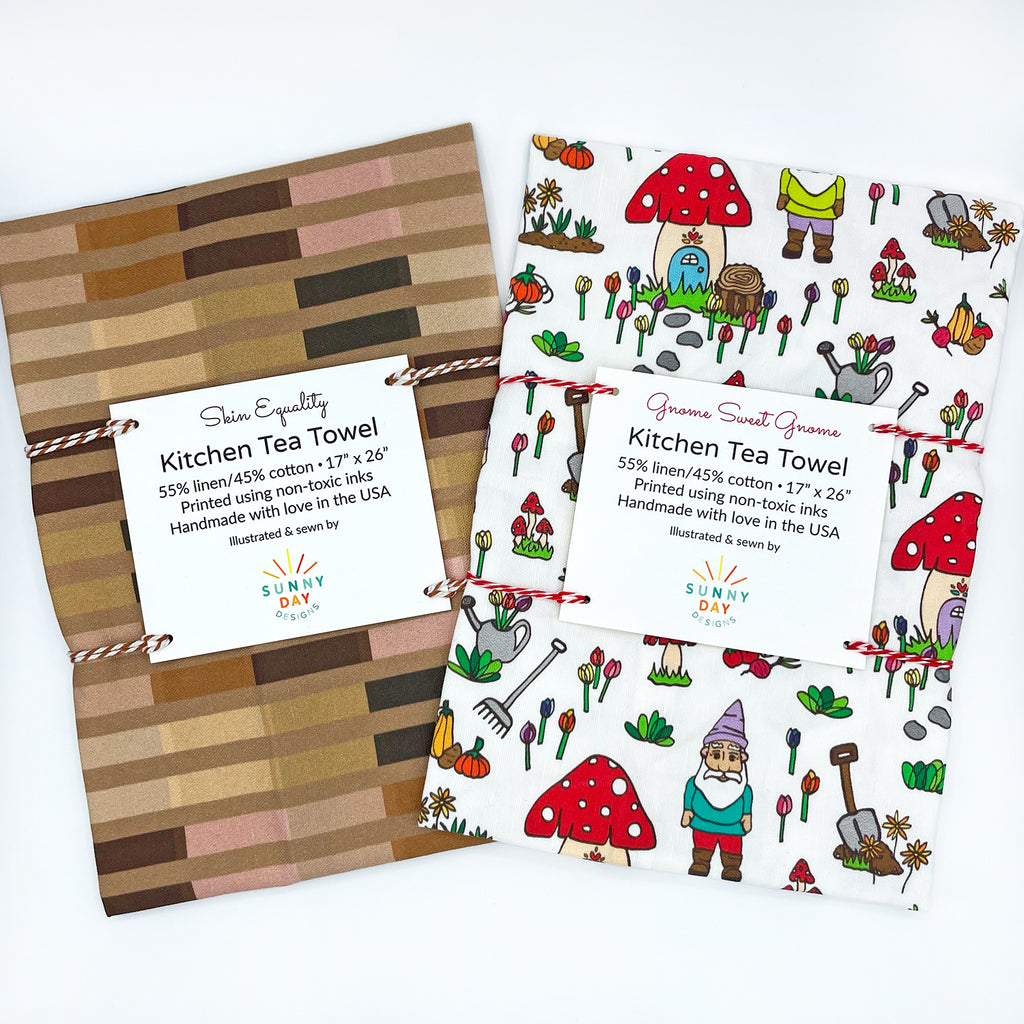 Set of two packaged kitchen tea towels by Sunny Day Designs. The first, Skin Equality, features various skin tone stripes, while the second, Gnome Sweet Gnome, showcases a playful gnome and garden-themed design with mushrooms, flowers, and tools. Both tea towels are made with a blend of linen and cotton.