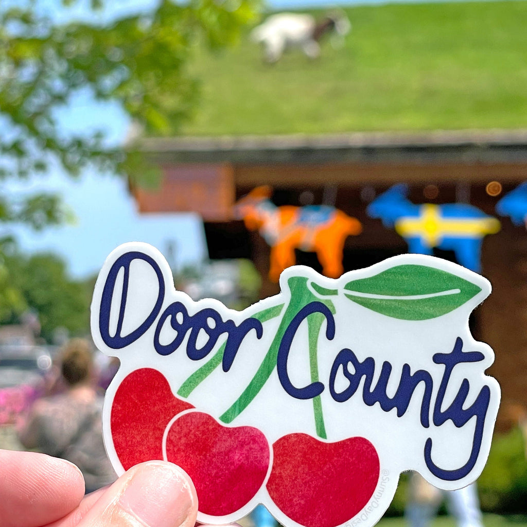 A Door County, Wisconsin, cherry-themed souvenir vinyl sticker is shown held in 1 hand in front of the popular Door County restaurant, Al Johnson’s Swedish Restaurant, where a white/brown goat can be seen in the background of the photo grazing on top of the grassy restaurant roof.