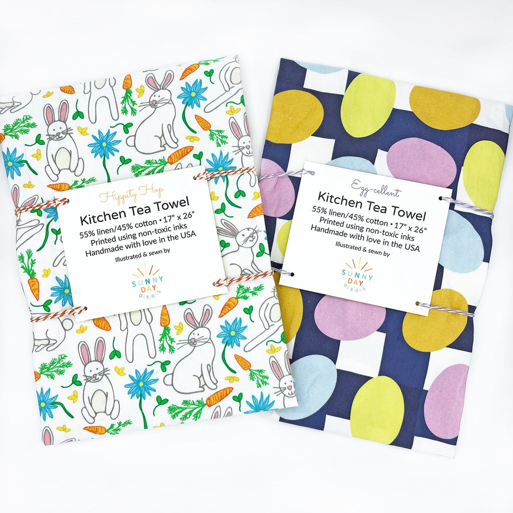 A set of 2 colorful Easter-themed printed kitchen tea towels by Sunny Day Designs are shown packaged on a white background, side-by-side. 1 features an Easter bunny print and the other features a minimalist Easter egg print