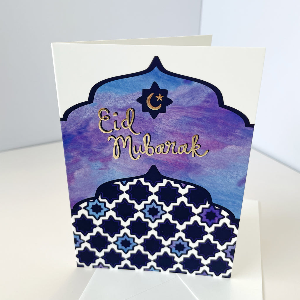 A colorful blue and purple watercolor Eid al-Fitr greeting card with metallic gold embossed "Eid Mubarak" text on cover is shown standing up with a white background and a corner in the background. This beautiful made in the USA Islamic greeting card is the perfect way to celebrate the end of Ramadan and send Eid greetings to Muslim friends, family & more.