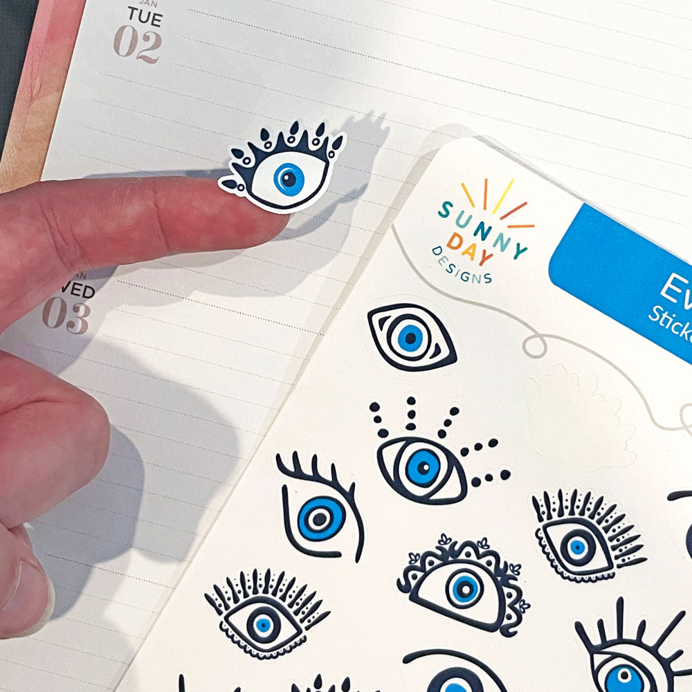 A pointer finger holds 1 blue evil eye-themed recycled paper sticker from an Evil Eye sticker sheet made by Sunny Day Designs hovers over a planner calendar page.