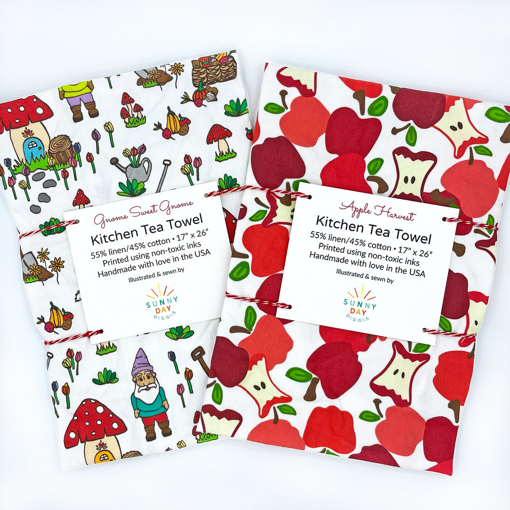 A 2-piece tea towel gift set  by Sunny Day Designs includes 1 multicolor whimsical garden gnome kitchen tea towel and 1 red apple harvest fruit-themed printed kitchen tea towel is shown packaged on a white background