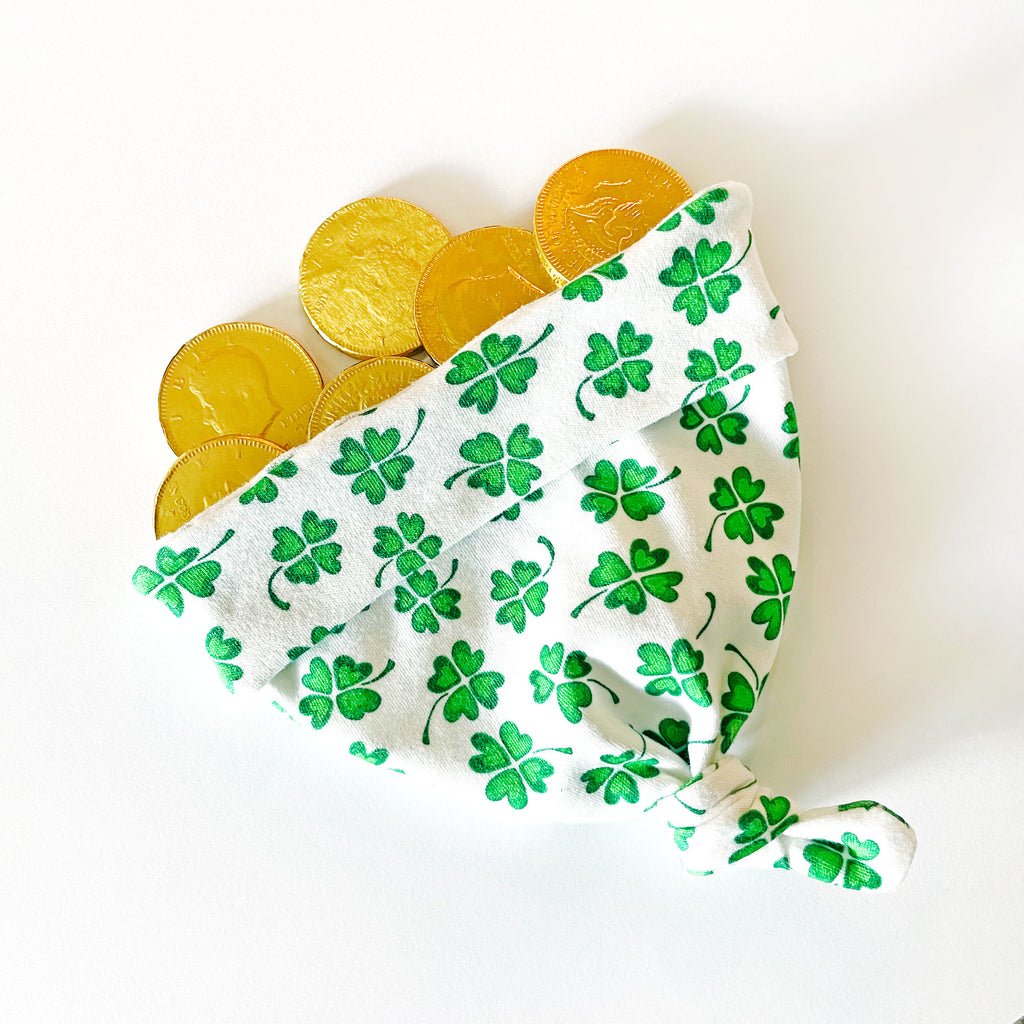 a green and white Irish-themed newborn baby hat is printed with shamrocks and is shown laying on a white background with golden coins coming out of it