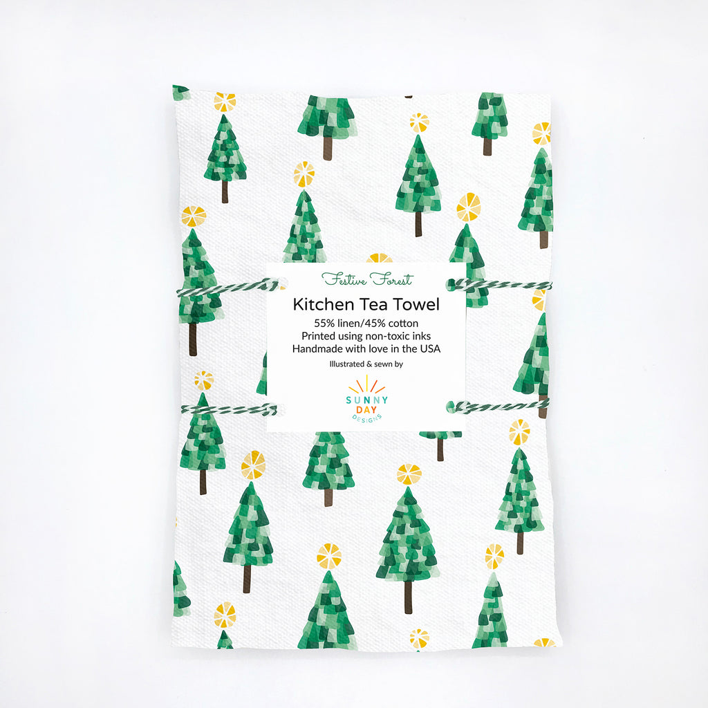 A handmade linen/cotton kitchen tea towel by Sunny Day Designs with a whimsical green, yellow and brown Christmas tree print is shown folded and packaged on a white background. These fun and eco-friendly printed holiday tea towels are joyfully made in the USA and are digitally printed with non-toxic inks.