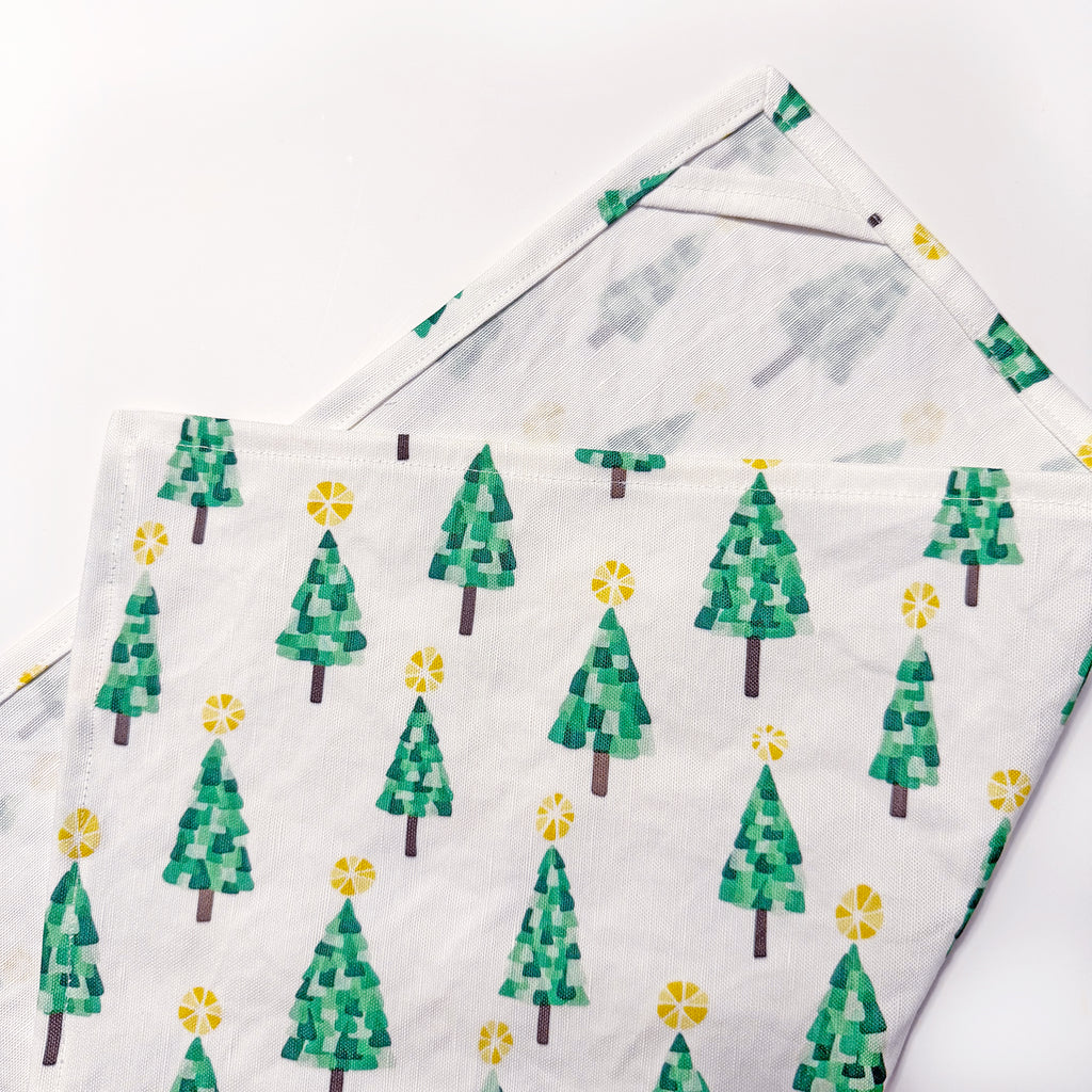 A colorful green, yellow, brown and white Christmas tree printed kitchen tea towel handmade by Sunny Day Designs is shown folded to highlight the printed outer side and also the seams around the outside of the dish towel, with the inside corner hanging loop visible.