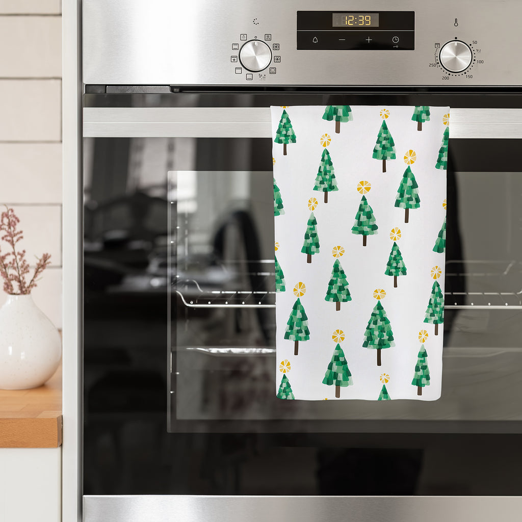 A colorful and festive Christmas tree patterned handmade kitchen tea towel by Sunny Day Designs is shown folded in half and hanging over the handle of a stainless steel oven in a kitchen. This "Festive Forest" printed dish towel is made in America in small batches from a linen/cotton fabric that is printed with exclusive print designs by Shelley Schmidt