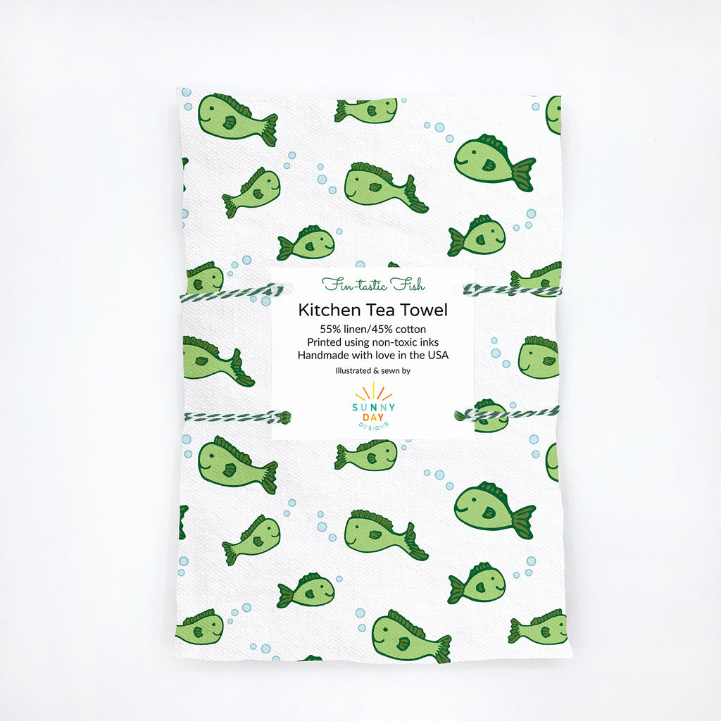 Folded kitchen tea towel featuring a playful green cartoon fish and bubble pattern on a white background, made from 55% linen and 45% cotton. Packaged with a white label that reads “Fin-tastic Fish Kitchen Tea Towel – 55% linen / 45% cotton, printed using non-toxic inks, handmade with love in the USA.” Minimalist flat lay on a white background with eco-friendly branding by Sunny Day Designs