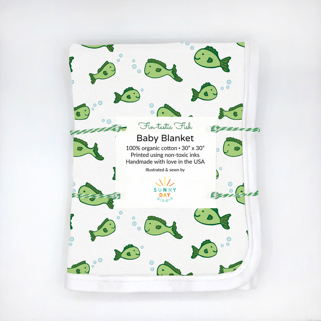 Folded organic cotton baby blanket featuring a playful green fish pattern with scattered light blue bubbles on a white background, packaged with a white label that reads “Fin-tastic Fish Baby Blanket – 100% organic cotton, 30” x 30”, printed using non-toxic inks, handmade in the USA.” Minimalist flat lay on a white background with eco-friendly branding by Sunny Day Designs.