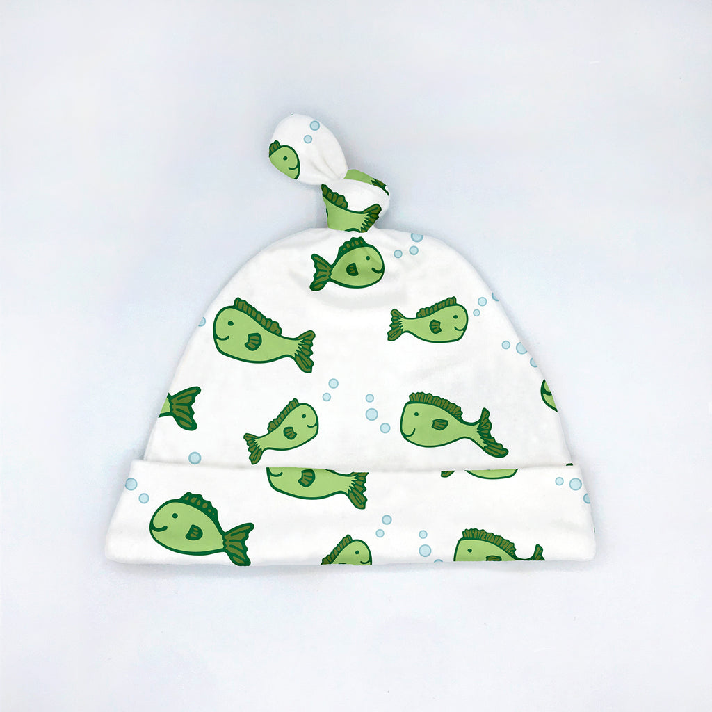 White baby hat with top knot featuring a repeating pattern of green cartoon fish and light blue bubbles, laid flat on a white background in a minimalist product display. Coordinates with the “Fin-tastic Fish” collection by Sunny Day Designs.