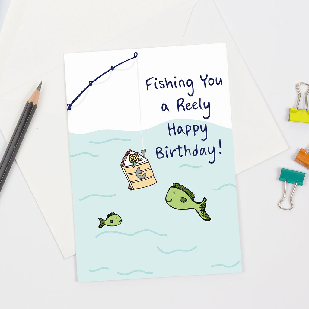 Greeting card featuring an illustrated fishing scene with two green cartoon fish and a slice of birthday cake hooked on a fishing line, with the pun message “Fishing You a Reely Happy Birthday!” in blue handwritten-style font. Displayed flat with an envelope and pencil on a white background.