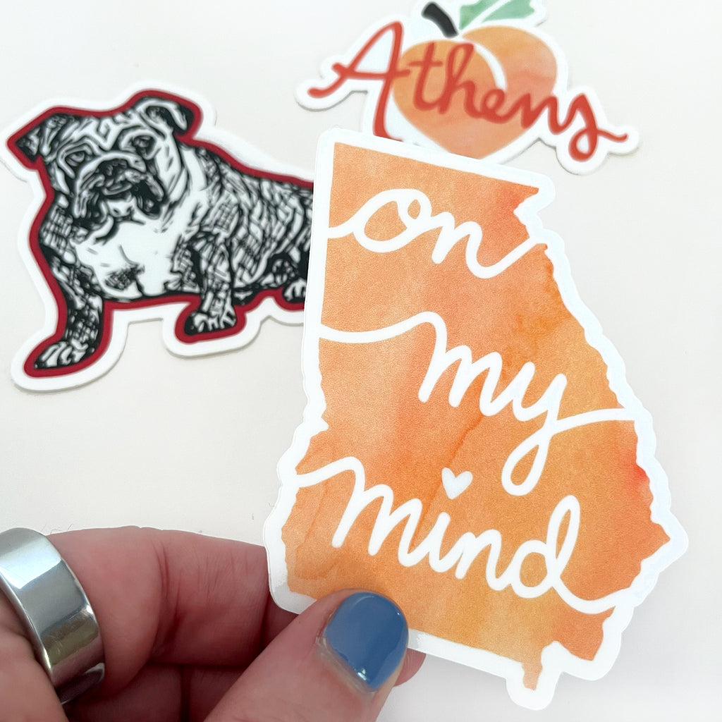 A Georgia state shaped watercolor peach vinyl sticker by Sunny Day Designs is shown held in a hand in front of a white background with 2 other University of Georgia Red Bulldog and Athens, GA peach stickers in the background. A perfect sticker gift set for any UGA alumni, graduate, student, or UGA football fan!