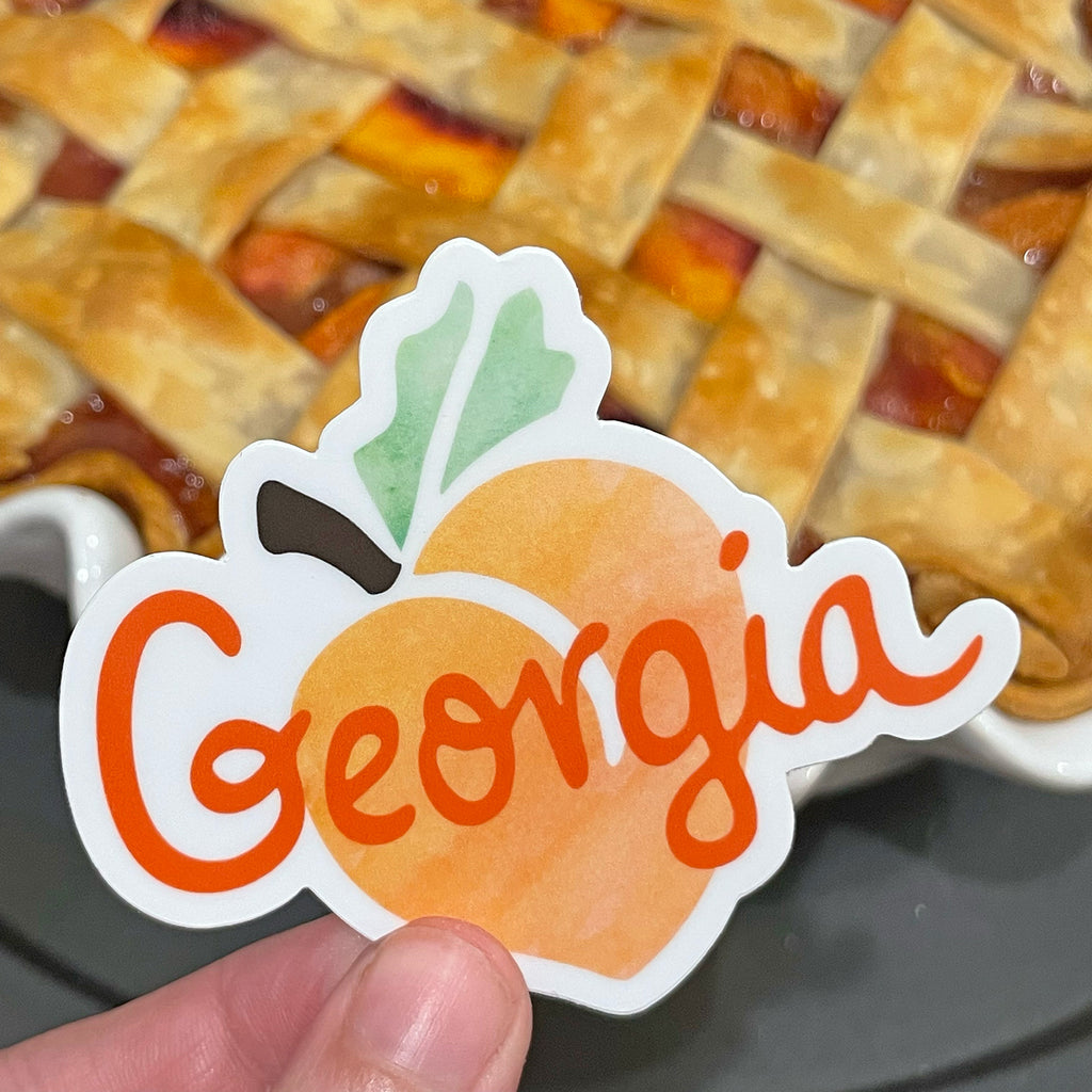 A photo of an orange, green and brown Georgia peach watercolor vinyl sticker by Sunny Day Designs is being held in 1 hand in front of a fully-baked peach pie with a lattice crust top.