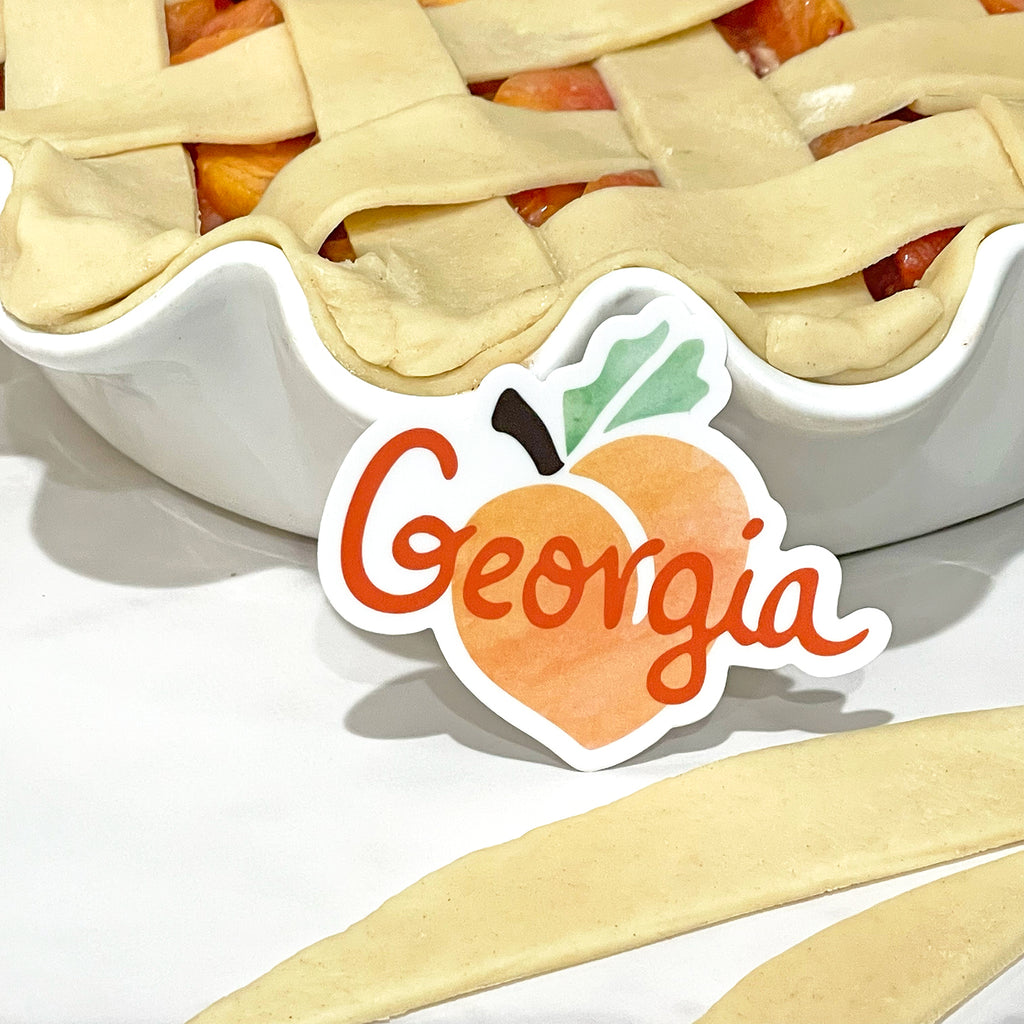 A small die cut vinyl sticker in the shape of an orange/green/brown/white Georgia peach design is resting against a pre-baked peach pie dish.