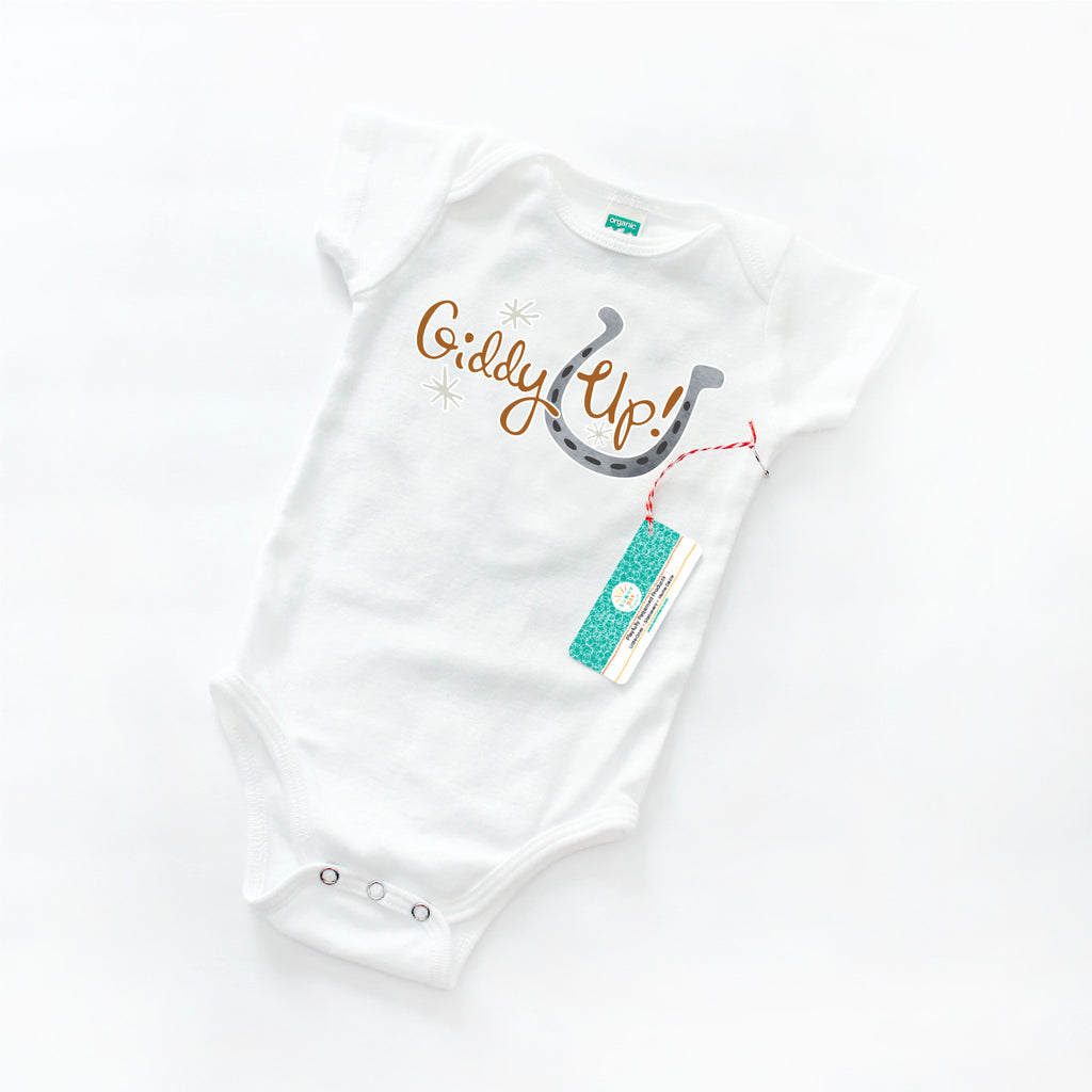 White organic cotton baby onesie printed with a horse-themed print design with brown "Giddy Up!" text, gray spur shapes, and a gray watercolor horseshoe design.