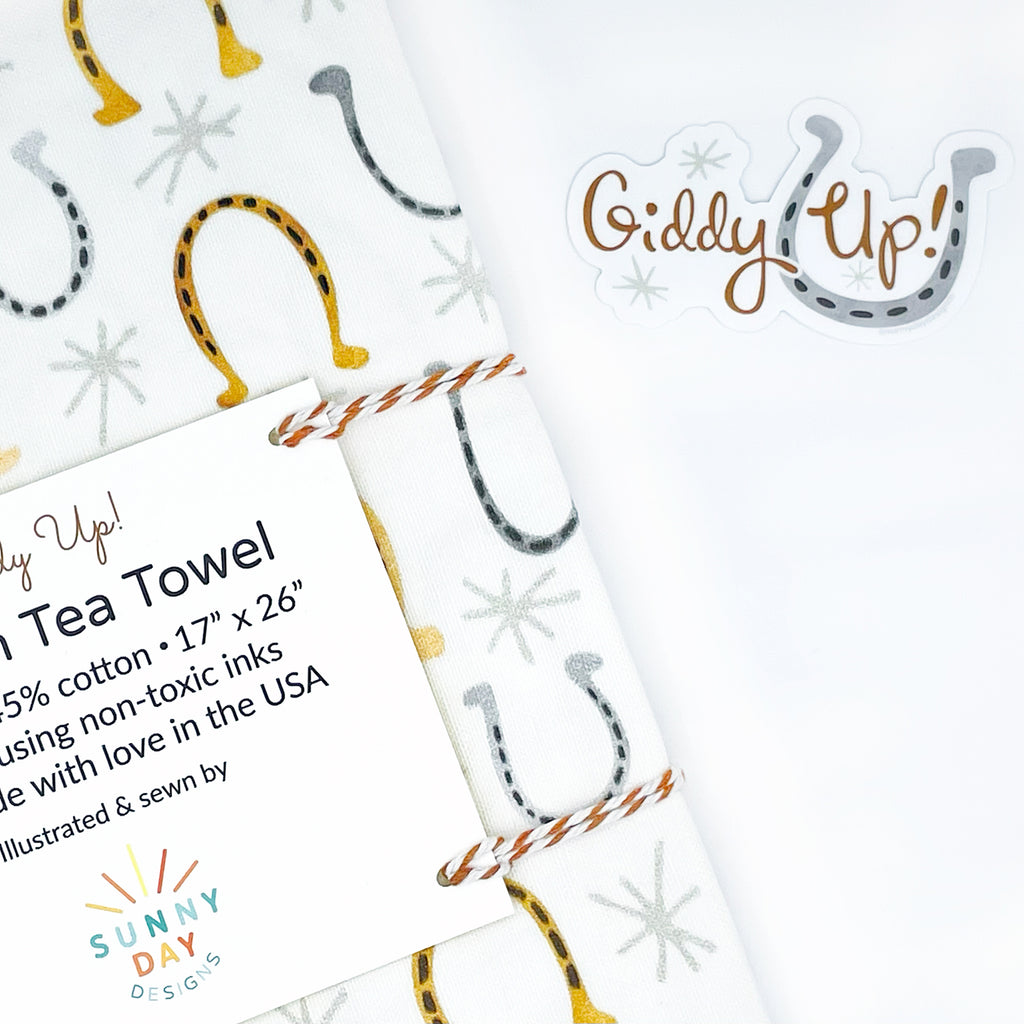 A Giddy Up! watercolor horseshoe printed tea towel by Sunny Day Designs is shown packaged with brown/white baker's twine & an informational card on a white background next to a horseshoe & equestrian themed vinyl sticker that says "Giddy Up!" in brown hand lettered text next to a gray watercolor horseshoe. Perfect for Kentucky Derby parties & horse lovers both the equestrian printed tea towel & the horse-themed vinyl sticker are joyfully made in the USA & were designed by Sunny Day Designs in Madison, WI.