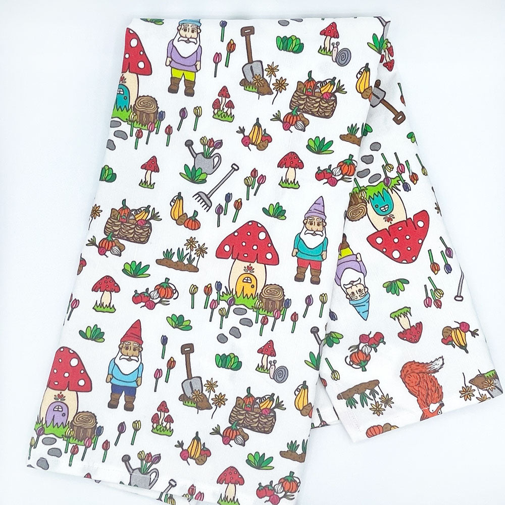 Folded Gnome Sweet Gnome kitchen tea towel featuring whimsical gnome-themed illustrations, including garden tools, mushrooms, and colorful flowers. The playful design is printed on a white background, perfect for adding charm to any kitchen.