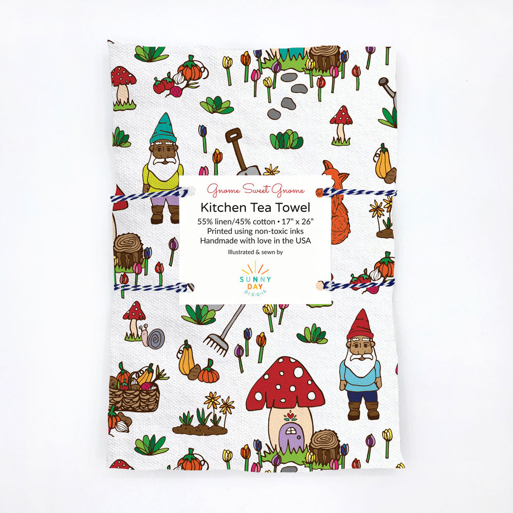 Packaged Gnome Sweet Gnome kitchen tea towel, with a label displaying its linen and cotton blend and handmade details. The design features gnomes, mushrooms, and garden tools, creating a fun and vibrant pattern on a white background.