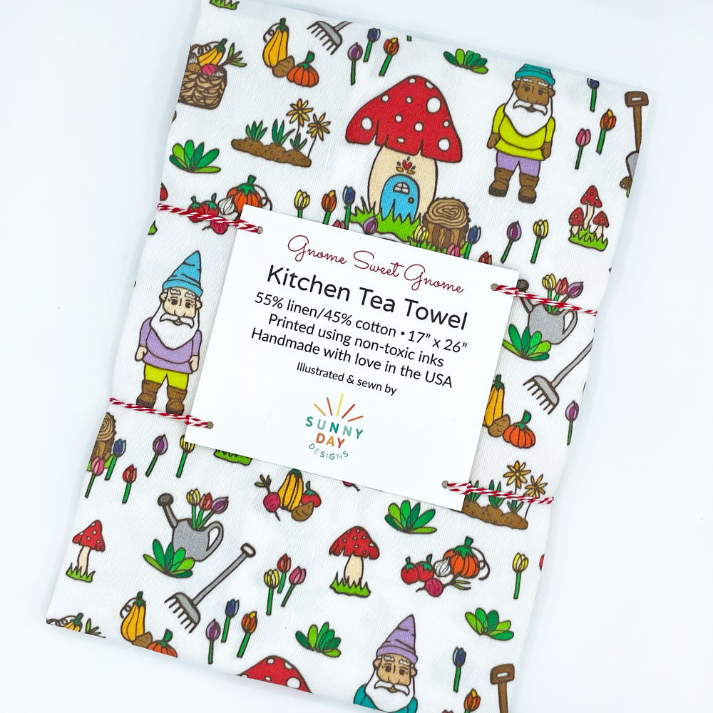 Close-up of the Gnome Sweet Gnome kitchen tea towel in its packaging. The towel showcases cheerful gnome characters, baskets of produce, and gardening tools, all set against a white backdrop. The tea towel is tied with red and white string, enhancing its gift-worthy presentation.