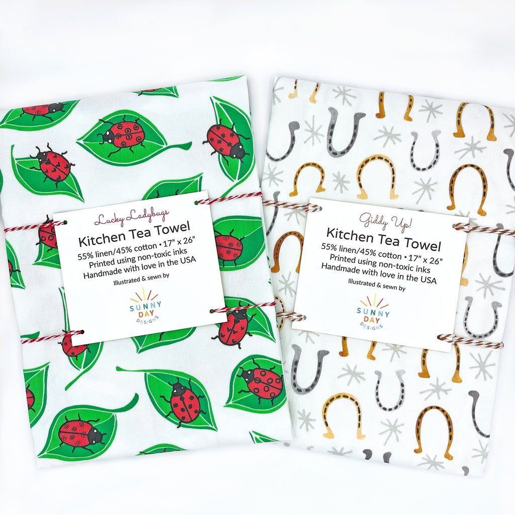 A set of 2 tea towels with a "Good Luck" theme are shown packaged and on top of a white background - included in the set are 2 tea towels featuring common lucky charms: 1 red/black/green ladybug printed tea towel and 1 gray and golden brown horseshoe printed tea towel