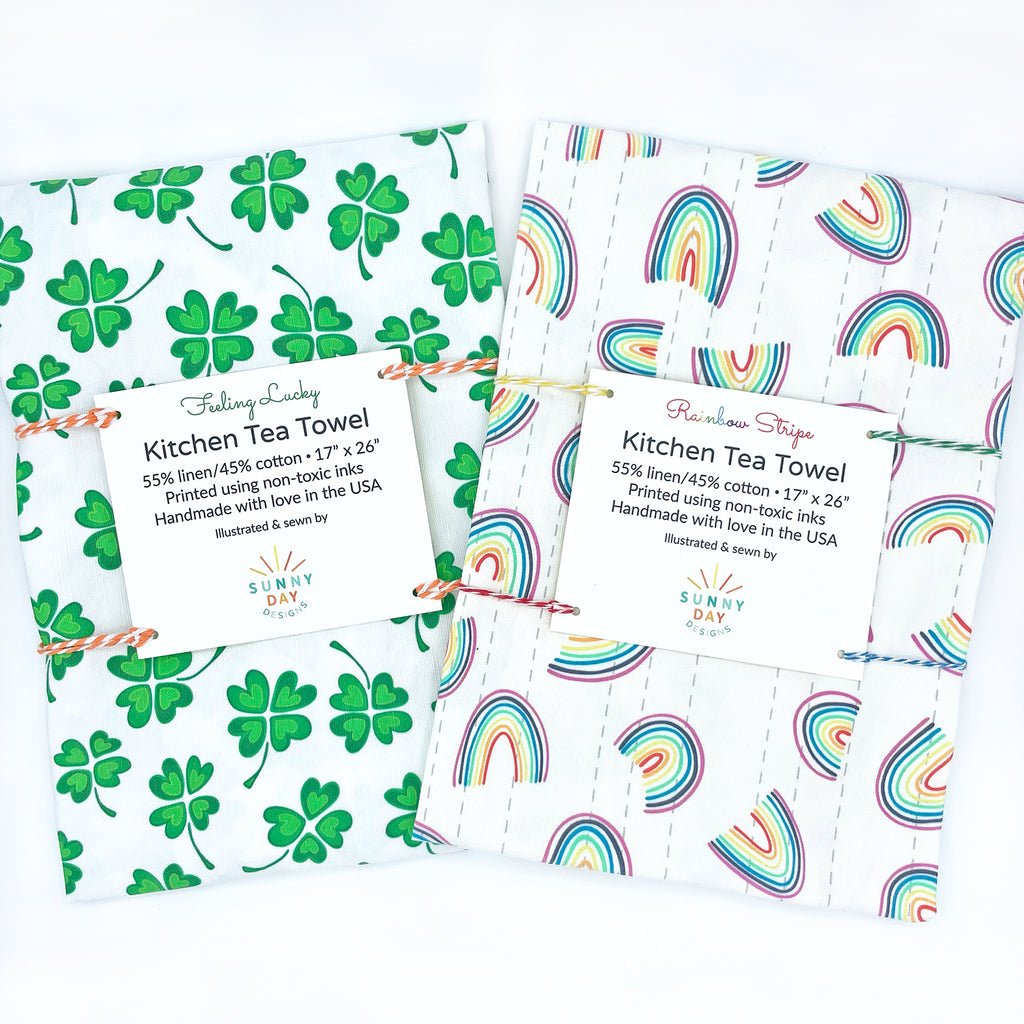 2 Packaged handmade linen/cotton tea towels are shown side-by-side on a white background as part of a St. Patrick's Day-themed gift set that includes 1 green four-leaf clover Irish-inspired tea towel and 1 rainbow themed tea towel.