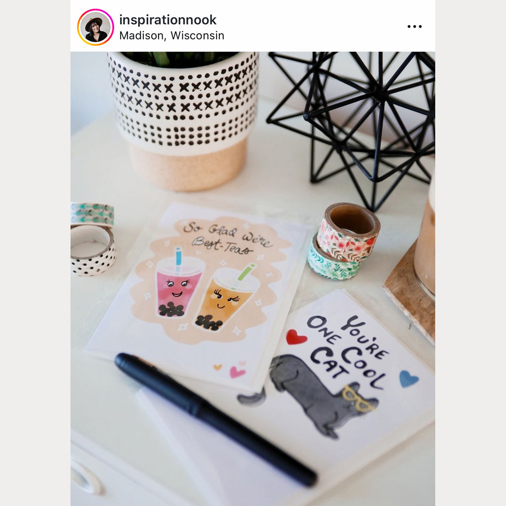 A cat-themed appreciation greeting card is shown on a white background next to a pencil and colorful binder clips - the cover of the greeting card shows a gray watercolor cat wearing yellow glasses, red, green, and blue hearts, and hand lettered black text that says "You're One Cool Cat"