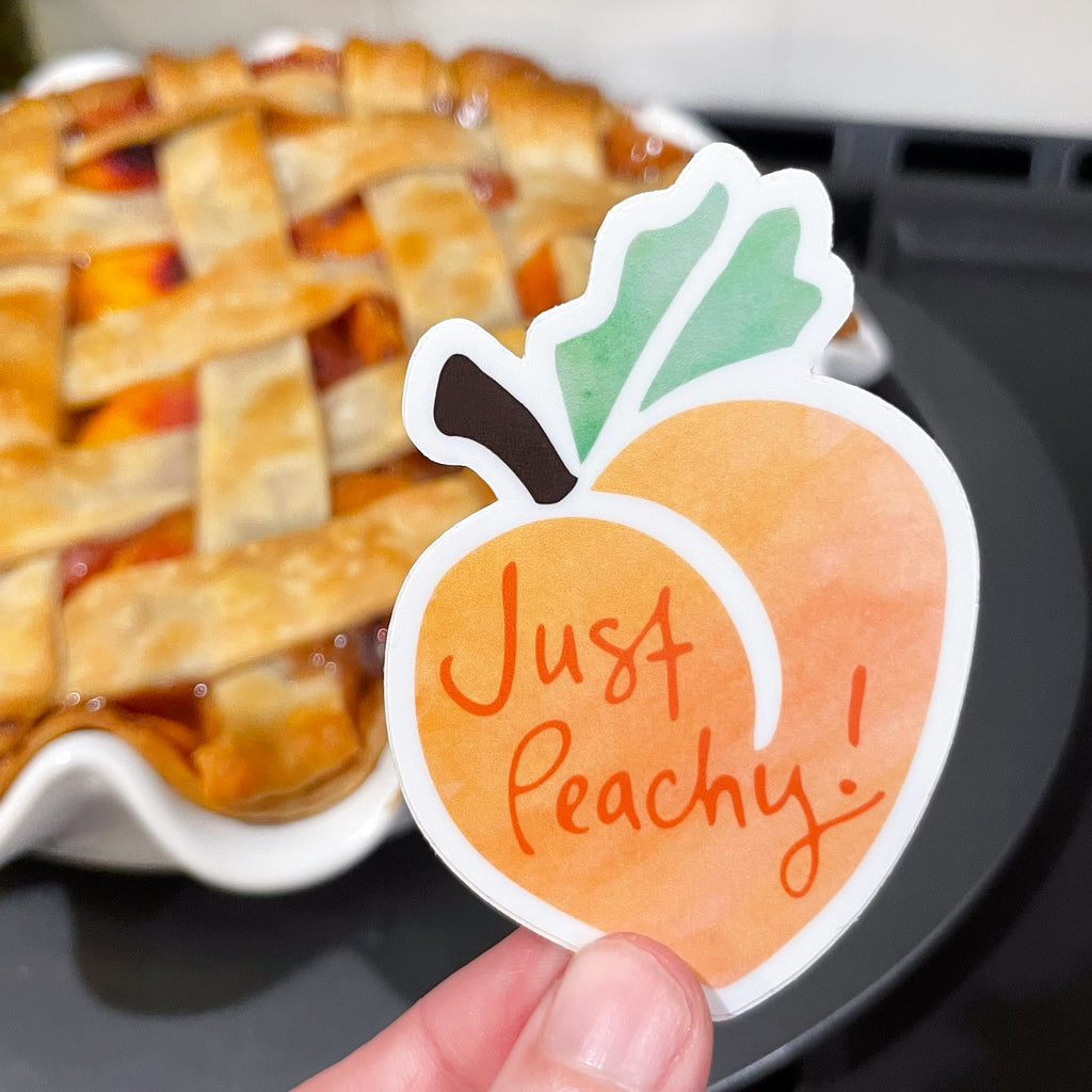 This close up image shows a hand holding an orange, green, and brown peach vinyl sticker that says "Just Peachy" by Sunny Day Designs. In the background you see a homemade peach pie with a lattice crust cooling on a kitchen stove.