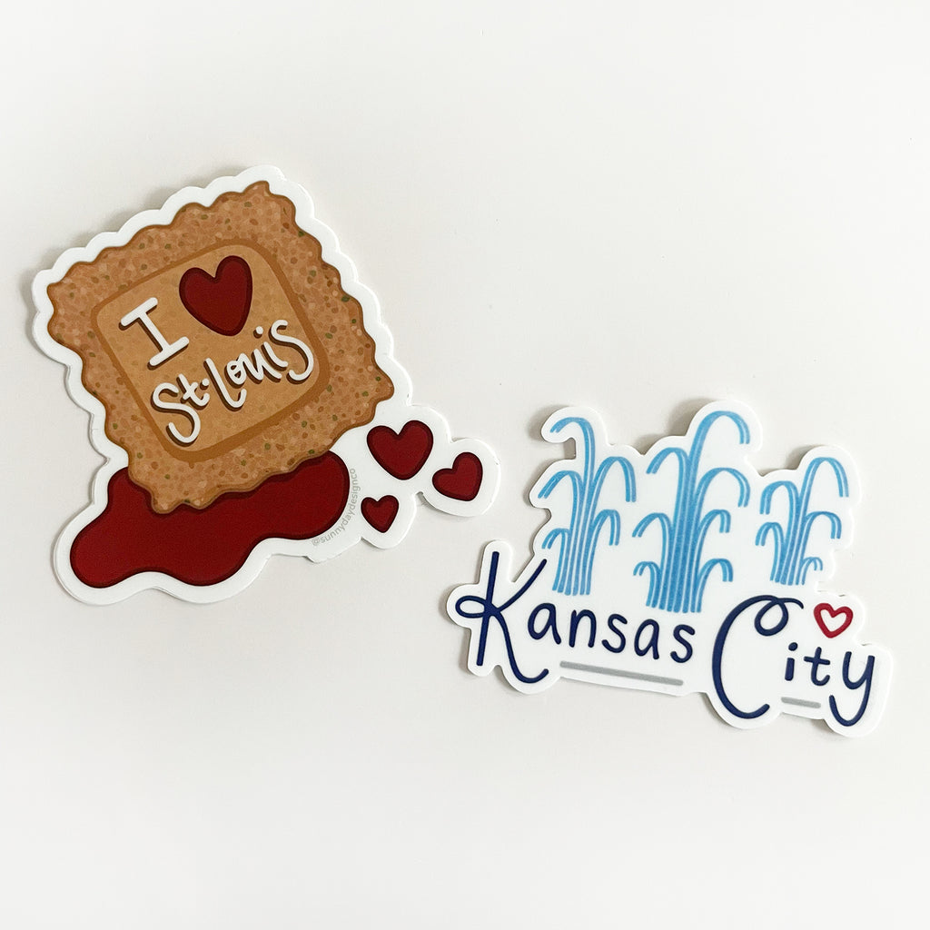 2 Missouri city-themed vinyl sticker designs by Sunny Day Designs are displayed on a white background in this image - An I Love St. Louis Toasted Ravioli-themed vinyl sticker is shown on a white background laying next to a Kansas City Fountain-themed vinyl sticker. Both of these colorful and cute Missouri stickers are made in the USA and designed by Sunny Day Designs in Madison, WI.