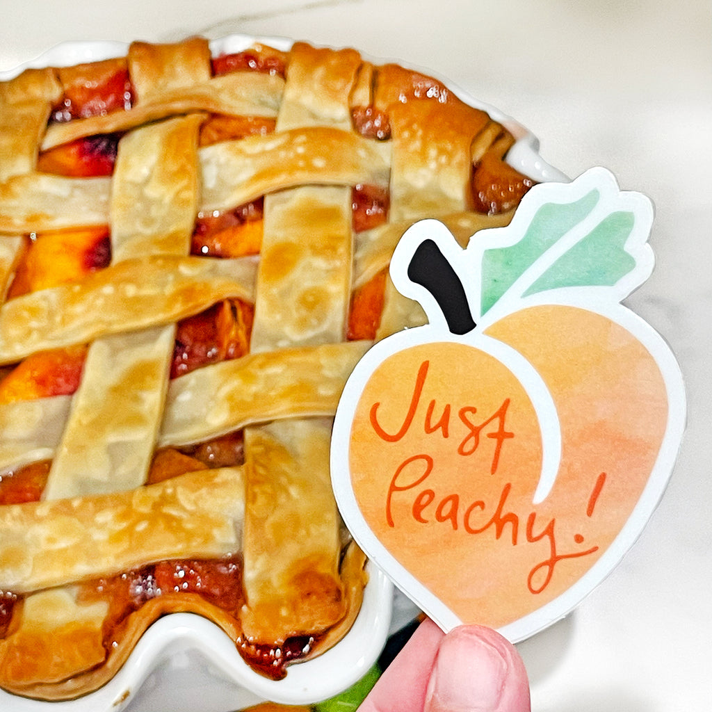 A sweet, orange, green, brown & white peach fruit-themed "Just Peachy" vinyl sticker by Sunny Day Designs is held in 2 fingers in front of a freshly baked peach pie with lattice crust