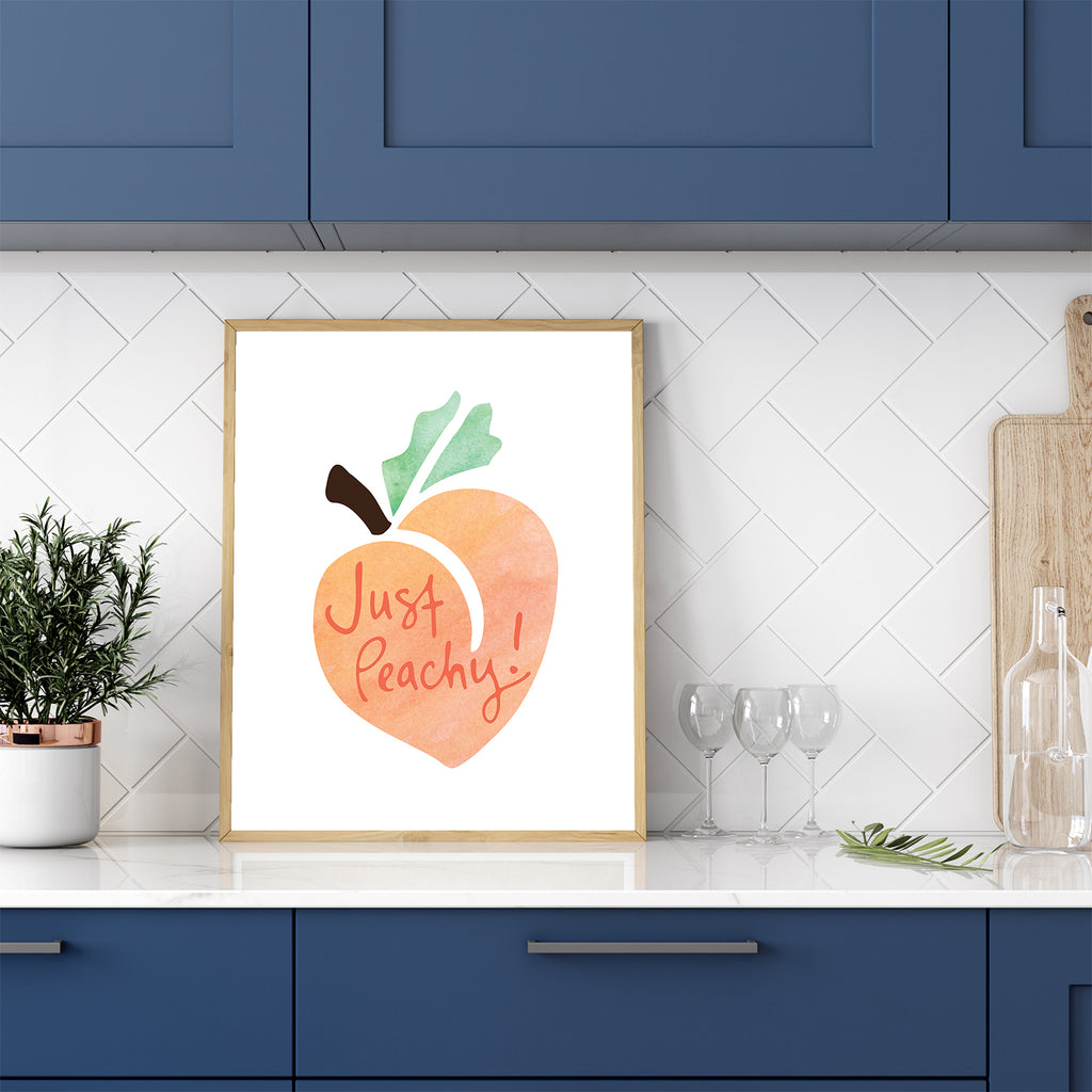 A fun framed wall art print (the Just Peachy 8x10 watercolor art print by Sunny Day Designs) is shown leaning against white subway tile on top of a white kitchen counter with blue cabinets. This colorful fruit-themed wall art features hand lettered "Just Peachy!" text on top of an orange, green and brown watercolor peach illustration and highlights the whimsical traditional southern saying, "Just peachy!"