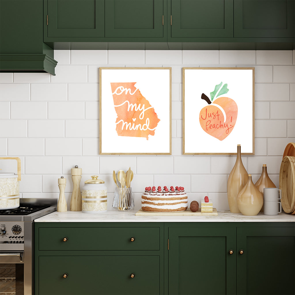 A beautiful and tastefully decorated kitchen with forest green cabinets and a white subway tile backsplash featuring 2 framed 8x10 sized art prints is shown with a beautifully decorated cake and other desserts on the countertop, alongside several decorative canisters, containers and utensils. Inside the 2 picture frames that are hung up on the backsplash are the Georgia On My Mind watercolor Georgia state-themed art print and the Just Peachy watercolor art print highlighting this fun southern saying.