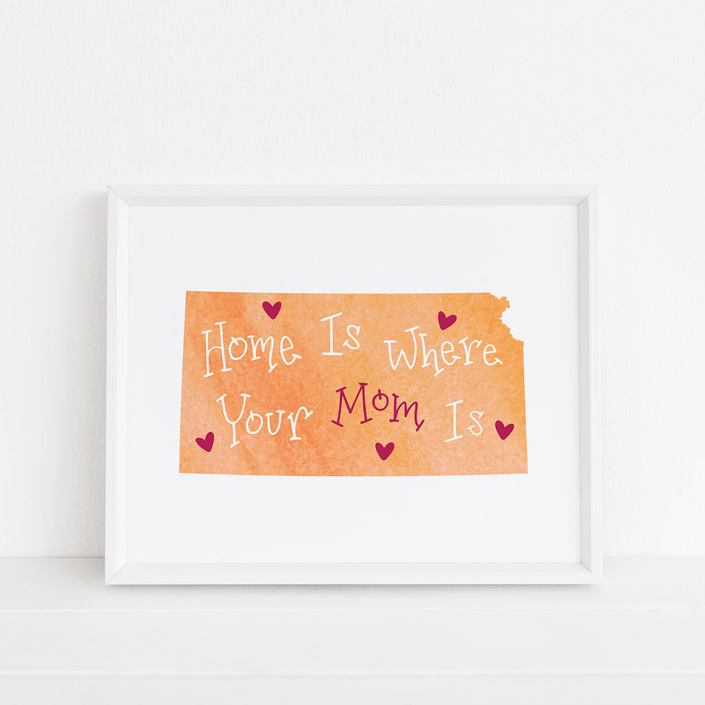 A framed "Kansas Mom" art print featuring a peachy orange watercolor silhouette of the state of Kansas with the hand-lettered phrase “Home Is Where Your Mom Is” in white and magenta lettering, accented with magenta hearts. Displayed in a white 8x10 frame on a white shelf, part of the Mother's Day and home state collection by Sunny Day Designs.
