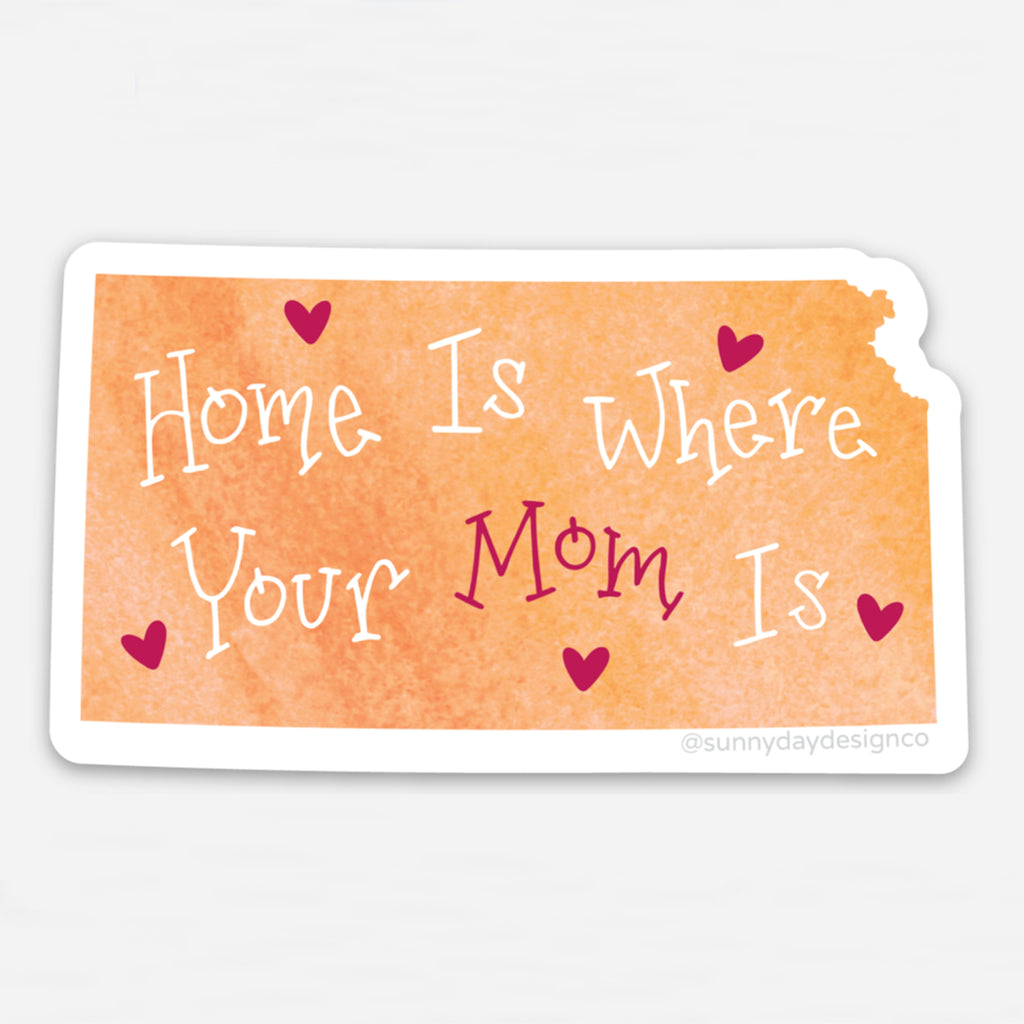 Vinyl sticker shaped like the state of Kansas with a watercolor-style orange background and white and magenta hand-lettered phrase “Home is Where Your Mom Is” with scattered heart accents. Die-cut with a white border, designed by Sunny Day Designs.