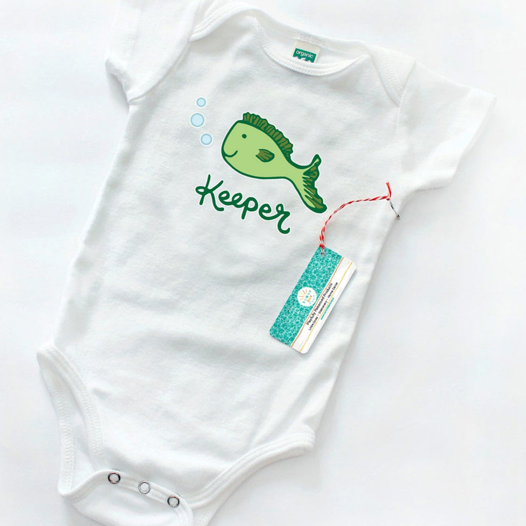 White baby onesie featuring a smiling green cartoon fish with light blue bubbles and the word “Keeper” in playful green script, displayed flat with a red and white hang tag labeled Sunny Day Designs. Minimalist flat lay on a white background with eco-friendly branding.