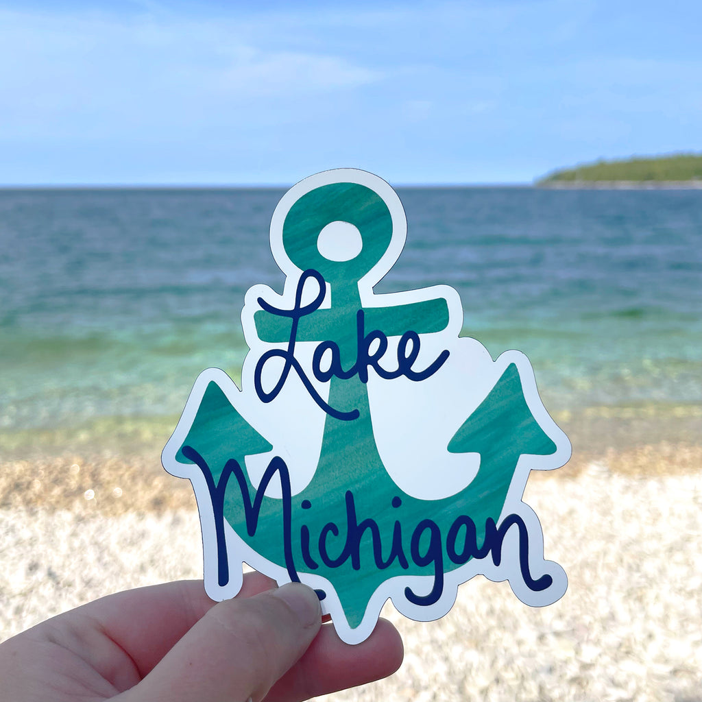 A cute Lake Michigan souvenir vinyl magnet (featuring a turquoise anchor and the words "Lake Michigan" in navy blue) is shown held in 1 hand in front of a beautiful beach and coastline on Washington Island, WI, which is on the shore of Lake Michigan.