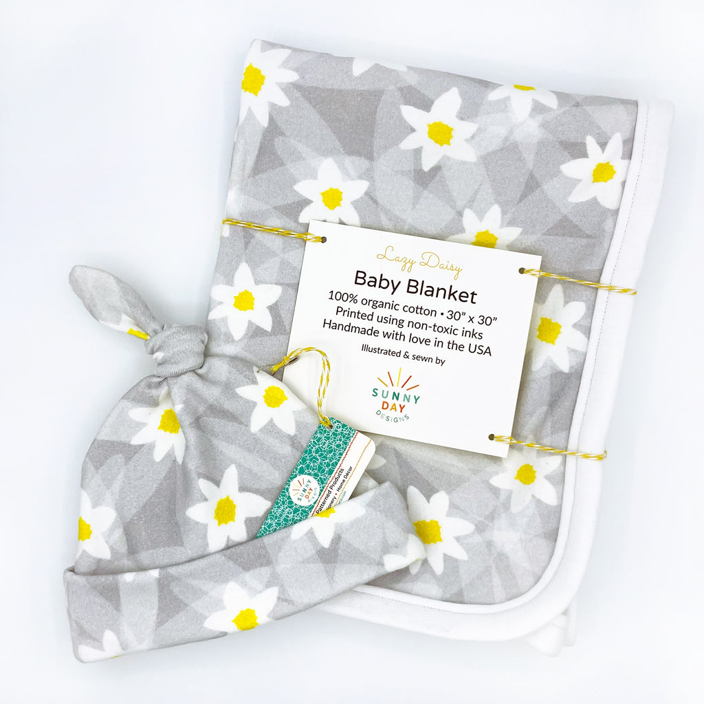 This image shows a cute grey, white & yellow abstract daisy floral printed organic cotton newborn baby hat and a coordinating Lazy Daisy printed receiving blanket, designed and handmade in the USA by Sunny Day Designs. Both the organic floral newborn hat and the organic floral baby blanket in this baby gift set are shown packaged on a white background