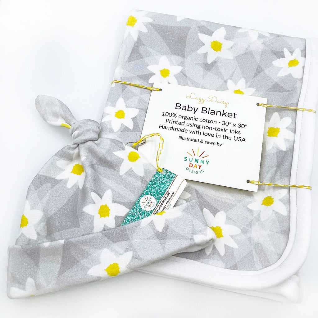 This image shows a tilted shot of an adorable grey, white & yellow abstract daisy printed organic cotton floral baby hat and a coordinating Lazy Daisy printed baby blanket, designed and handmade in the USA by Sunny Day Designs from organic cotton. Both the organic floral newborn hat and the organic floral baby blanket in this baby gift set are shown packaged on a white background