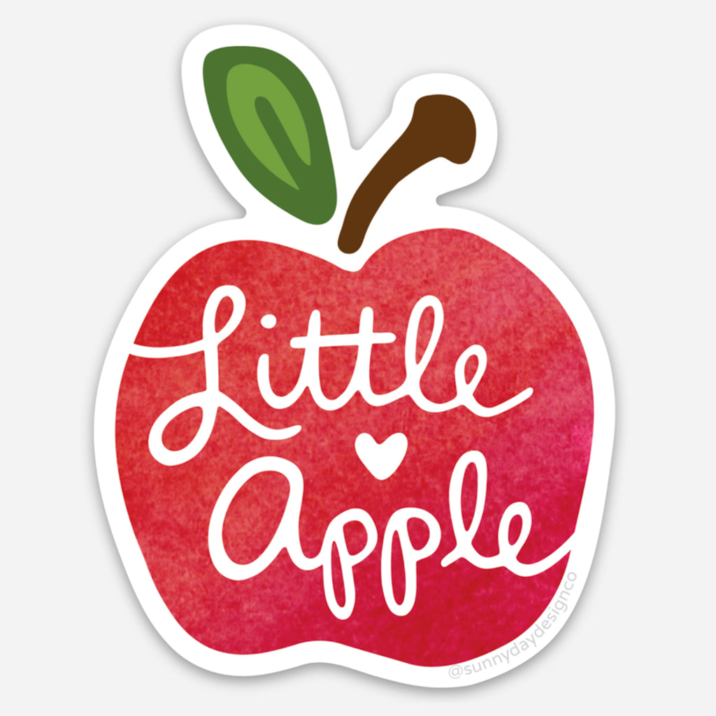 Vinyl sticker shaped like a red apple with a green leaf & a brown stem, featuring the hand-lettered phrase “Little Apple” in white script with a heart accent. Die-cut with a white border, inspired by the college city of Manhattan, Kansas, and designed by Sunny Day Designs.