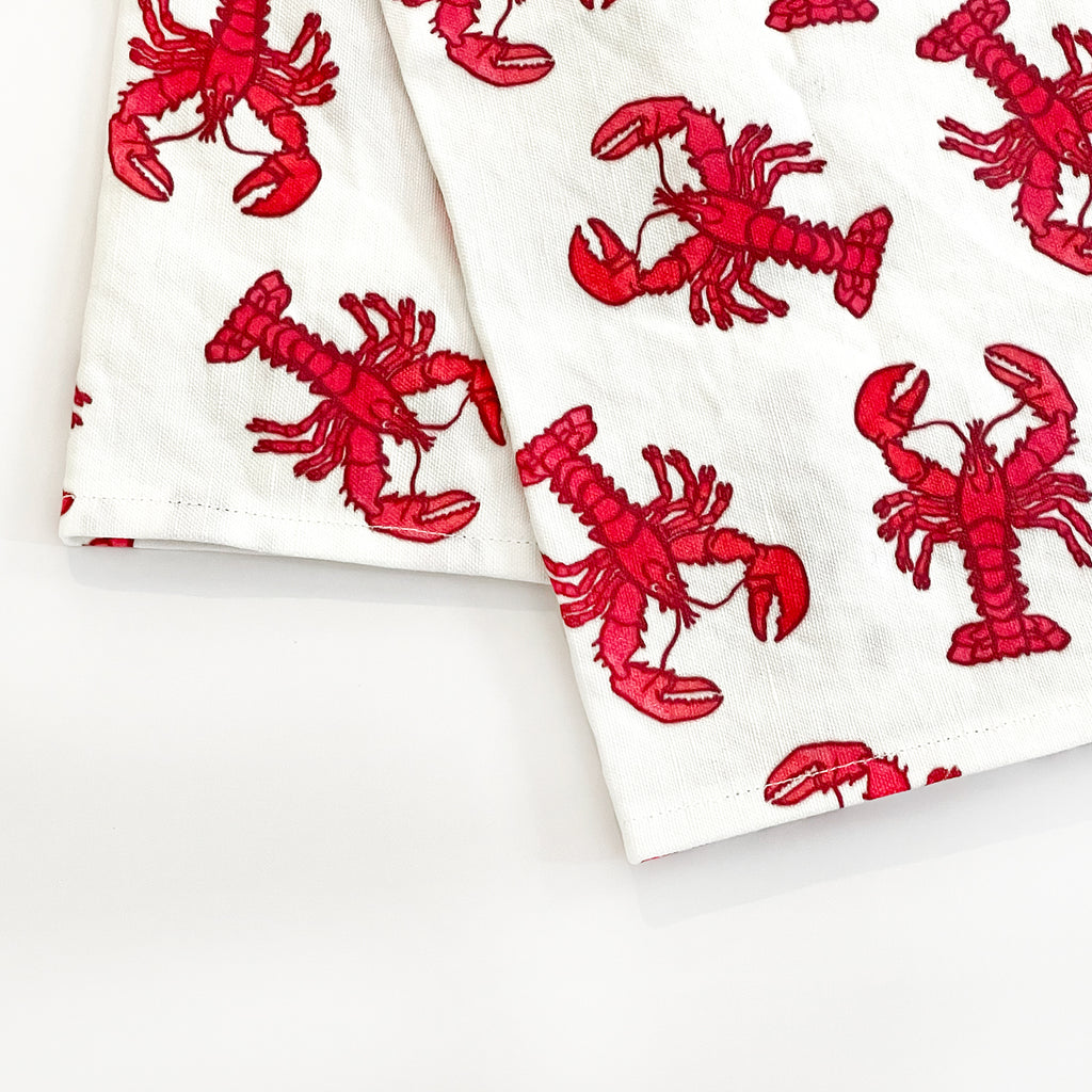 Close-up of a folded Lobster Tea Towel featuring vibrant red lobsters printed on a white background. The bold design adds a nautical touch to kitchen decor, perfect for seafood lovers.
