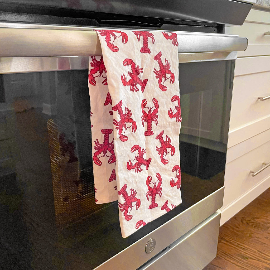Lobster Tea Towel hanging from a stainless steel oven door handle in a modern white kitchen. The towel’s striking red lobster design provides a bold pop of color and style to the space.