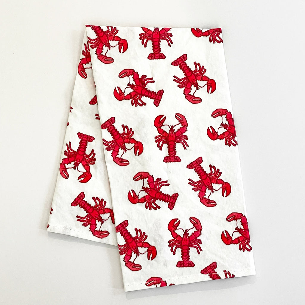 Folded Lobster Tea Towel with red lobsters printed in a repeating pattern across the white fabric. The towel is compact and adds a playful, coastal theme to any kitchen or dining setting.