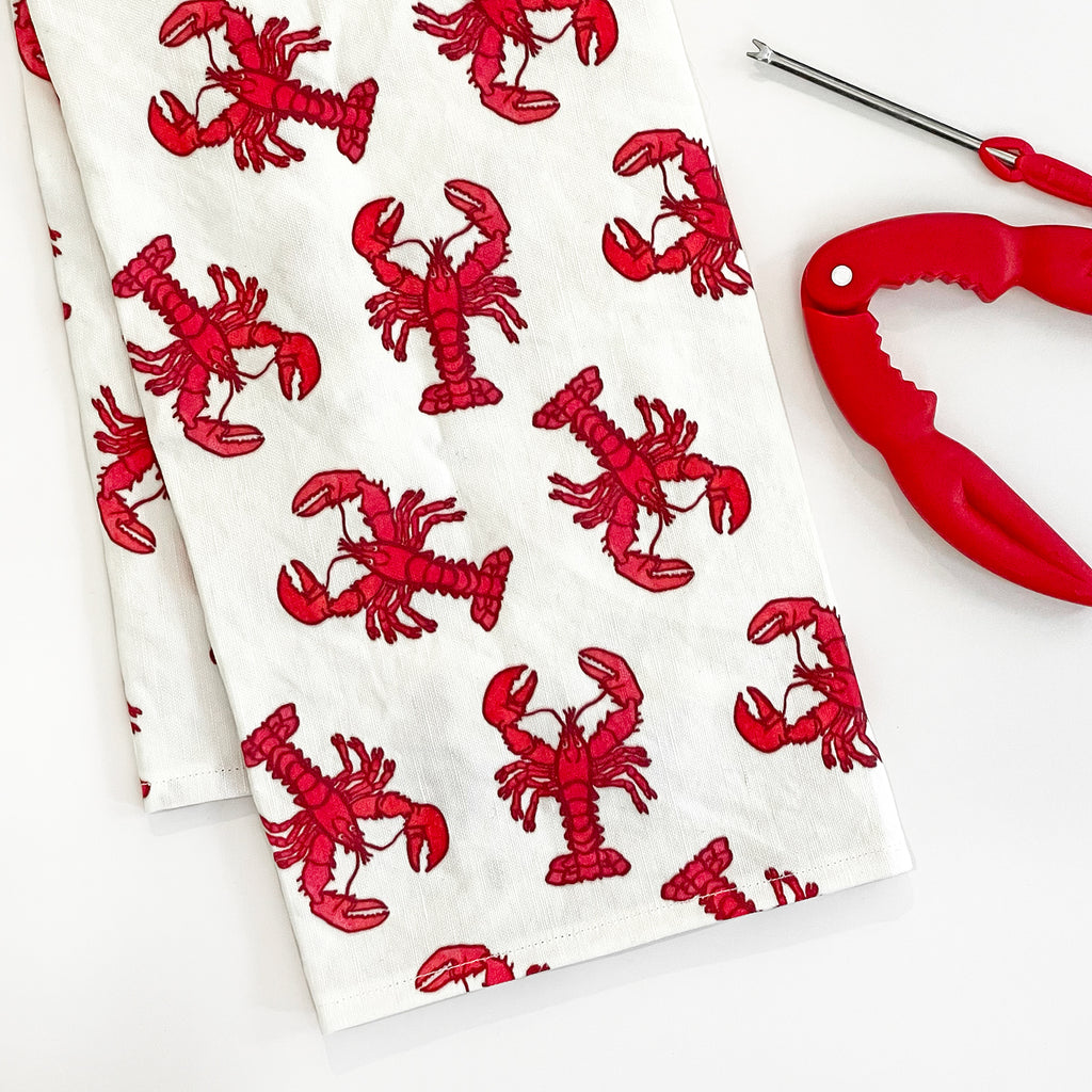 Lobster Tea Towel folded alongside a red lobster cracker tool, creating a cohesive lobster-themed set. The bright red lobsters on the white towel stand out as a fun and practical kitchen accessory.