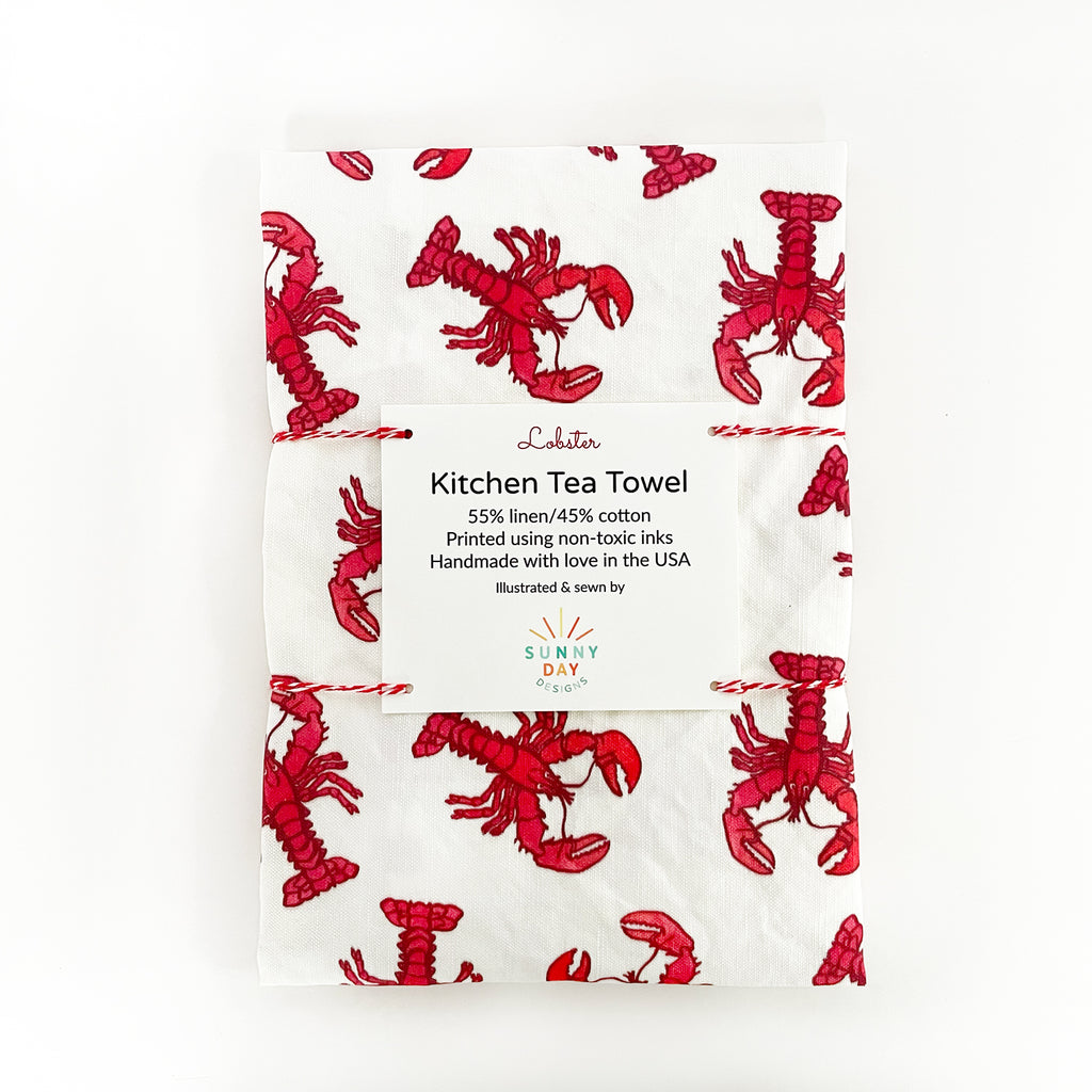 A white and red watercolor lobster patterned linen/cotton handmade tea towel by Sunny Day Designs is shown folded and tied with red/white baker's twine on top of a white background.